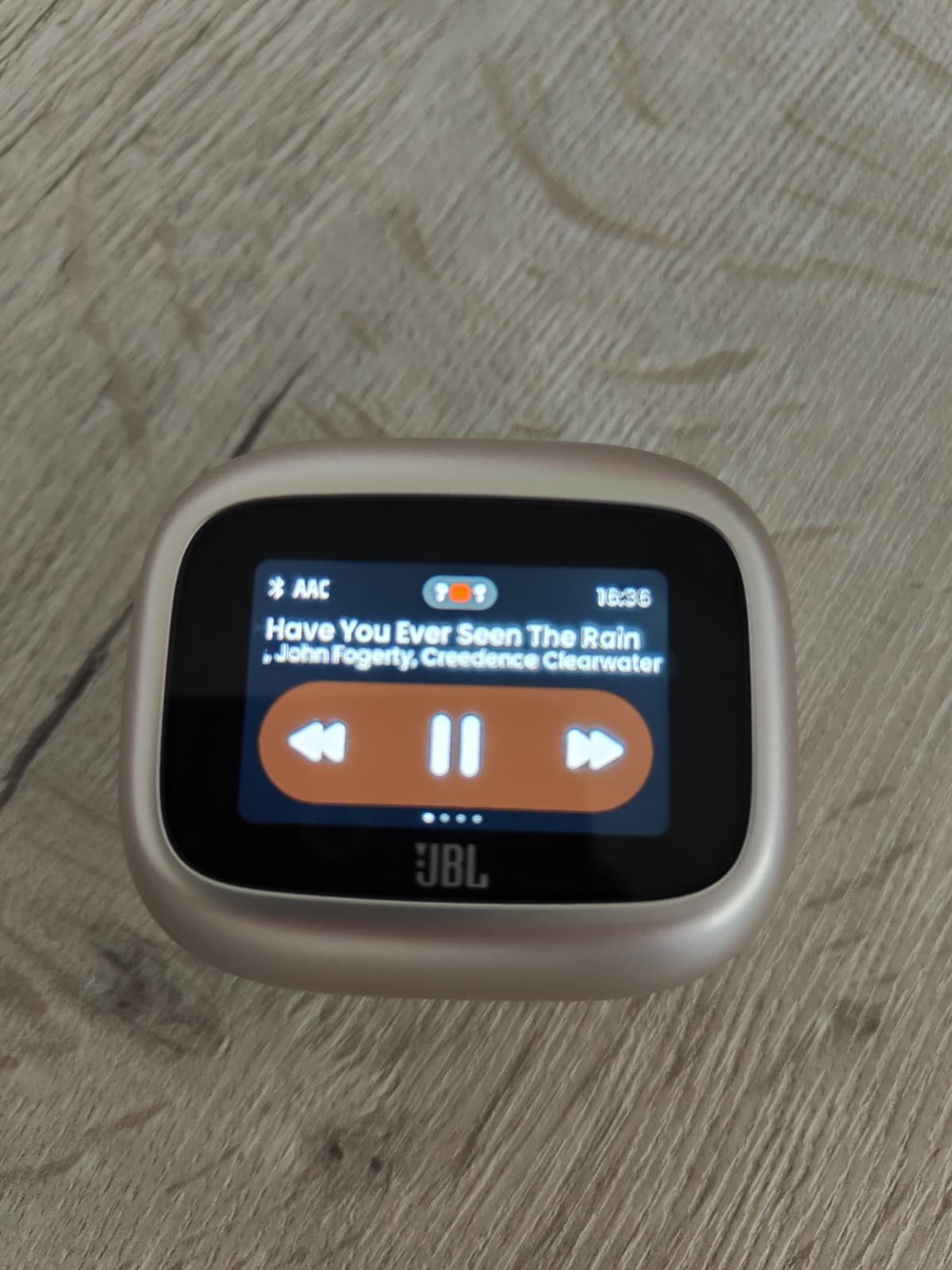 Scherm van de JBL Live Beam 4 case met het nummer dat speelt, de knoppen voor pauze en overslaan, en linksboven AAC